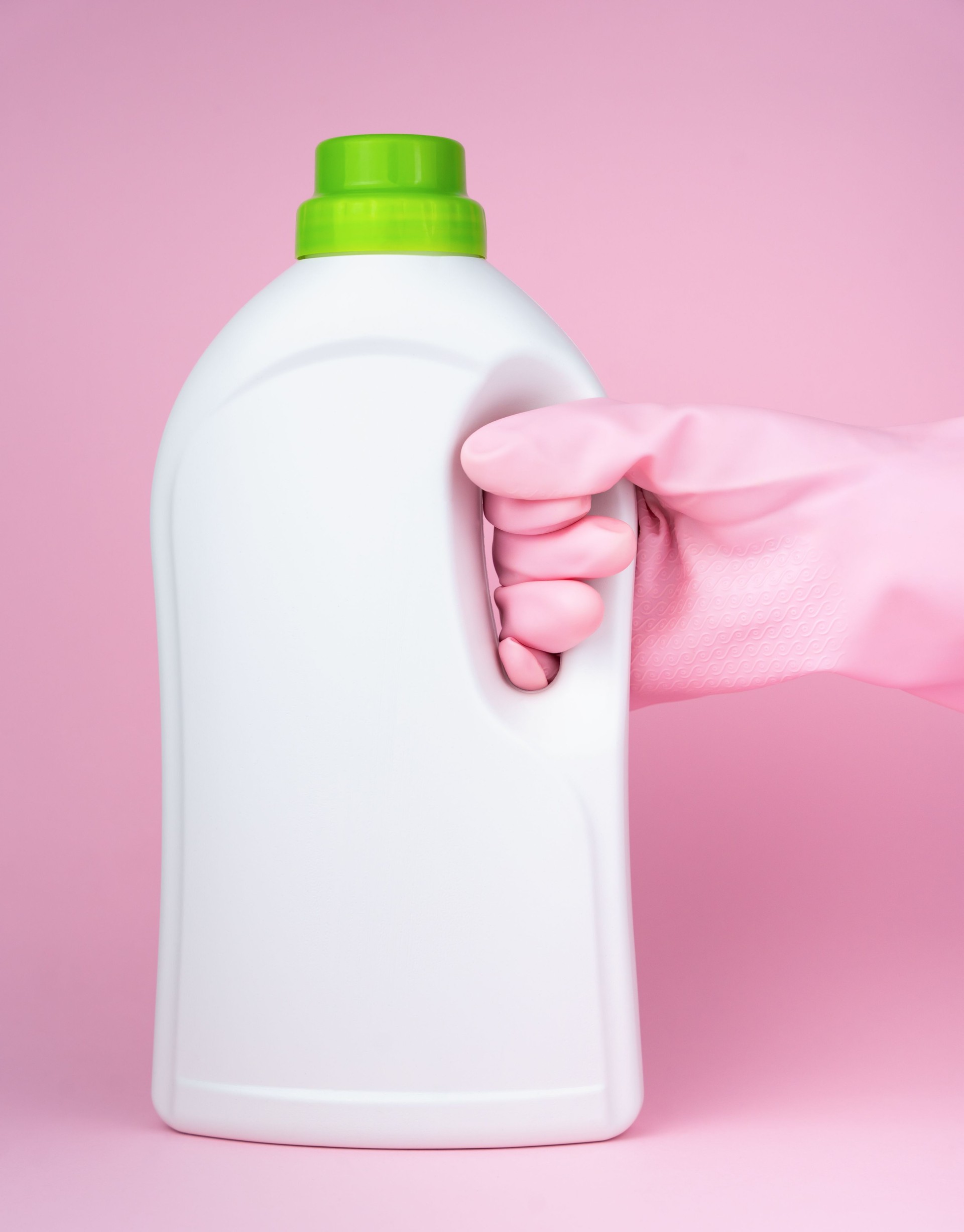 A hand in a pink rubber glove holds a bottle of laundry conditioner, softener bottle, liquid laundry detergent on a pink background. Close-up. Selective focus.